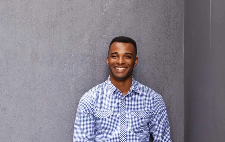 Mann i bermudashorts og skjorte som lener seg mot grå vegg