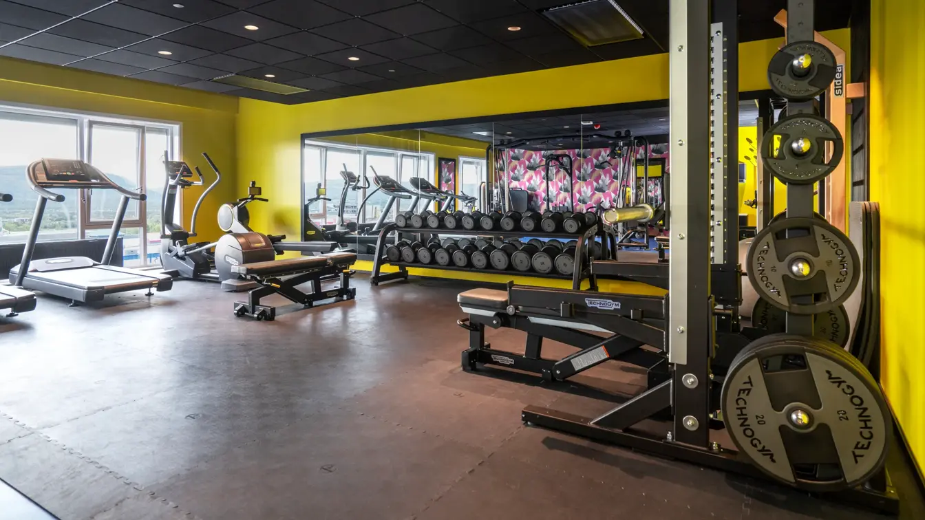 Svart gulv, gule vegger, veggspeil, diverse treningsapparater. 
