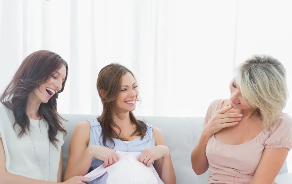 Cheerful pregnant woman at baby shower with her friends and looking at baby clothes at home