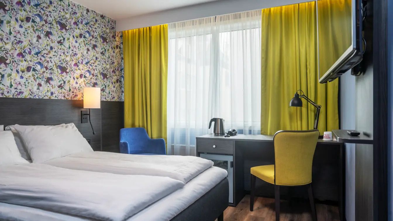 Gule gardiner, parkettgulv, gul stol, blomstertapet vegg, vindu, skrivebordpult, TV, dobbeltseng