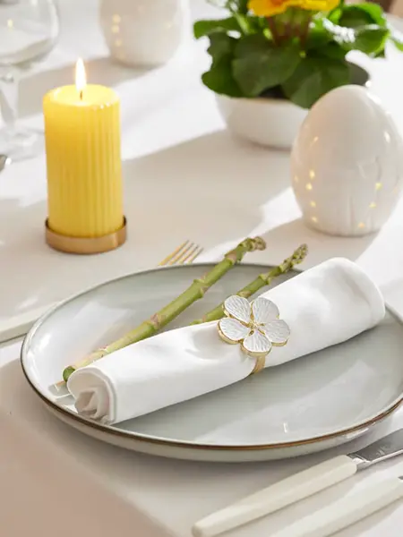 Kuvert på bord med hvit duk, gult kubbelys, pynteegg, blomst, tallerken med hvit serviett og serviettring.