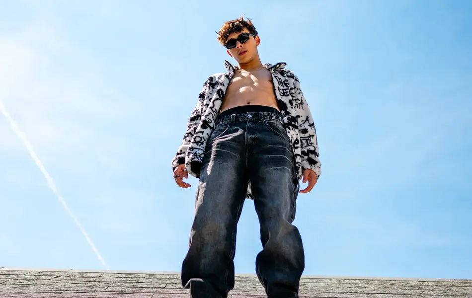 Mann med baggy jeans og bar overkropp under åpen hakke poserer utendørs, sett litt underfra 