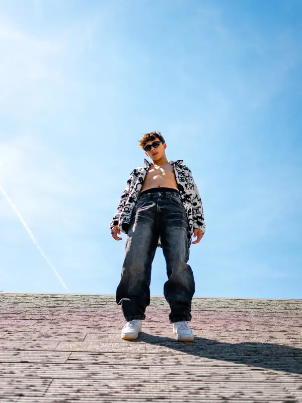 Mann med baggy jeans og bar overkropp under åpen hakke poserer utendørs, sett litt underfra 