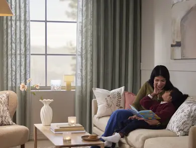Gardiner ved et vindu i en stue, med kvinne og barn i sofaen.