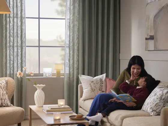 Gardiner ved et vindu i en stue, med kvinne og barn i sofaen.