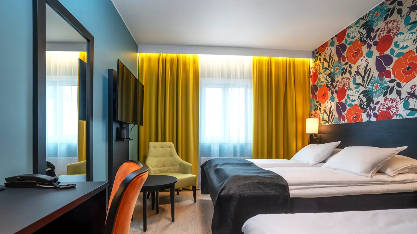 Blomstrete tapet, et stort vindu fra gulv til tak, gule gardiner, parkettgulv, tre enkle senger, svart skrivebord, en gul stol og en oransje stol. Et stort speil hengende på veggen.