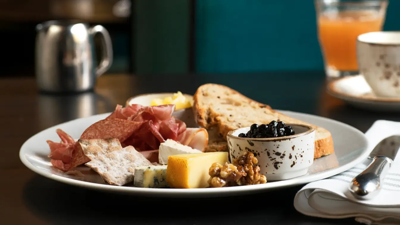 Frokosttallerken med ulike oster, syltetøy, nøtter, spekemat, knekkebrød og brødskive.