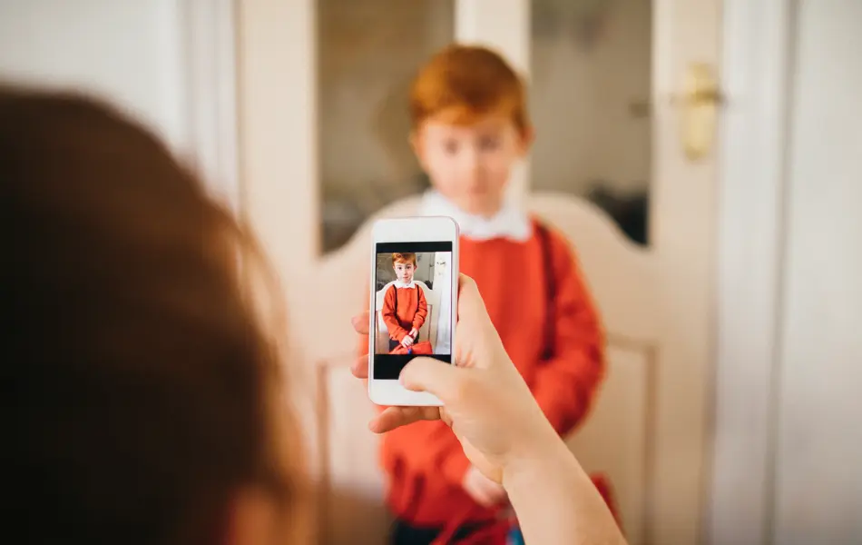 Little boy dressed for his first day back to school, having his photo taken by his Mother on her smartphone.