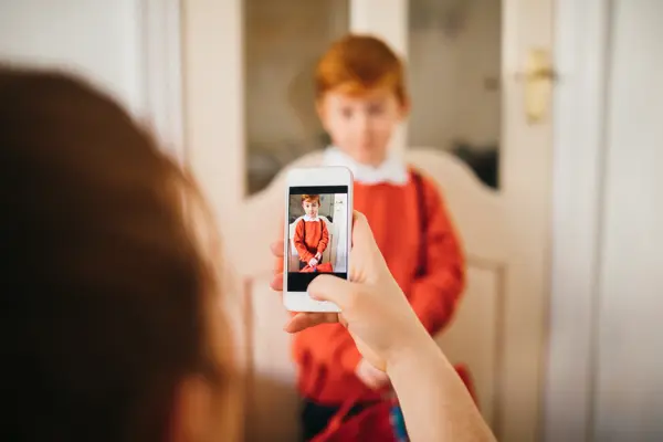 Little boy dressed for his first day back to school, having his photo taken by his Mother on her smartphone.