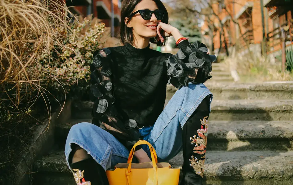 Dame sitter på trapp utendørs iført cowboyboots, en topp med transparante partier, jeans og solbriller. 