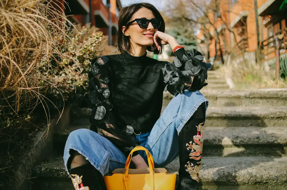 Dame sitter på trapp utendørs iført cowboyboots, en topp med transparante partier, jeans og solbriller. 