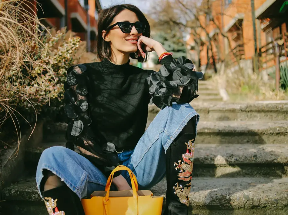 Dame sitter på trapp utendørs iført cowboyboots, en topp med transparante partier, jeans og solbriller. 