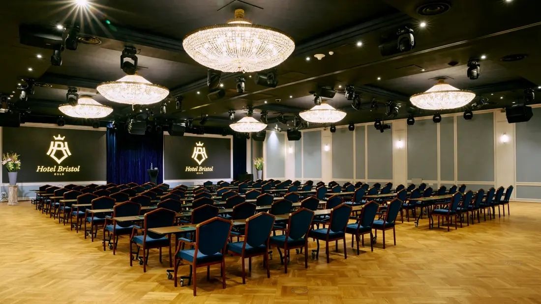 Møterom med klasseromsoppsett til 100 personer. Blå stoler, 6 store lysekroner i taket og to store skjermer foran. 