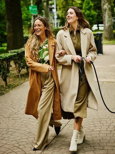 To damer i brune trenchkåper går hånd i hånd og ler på gaten 