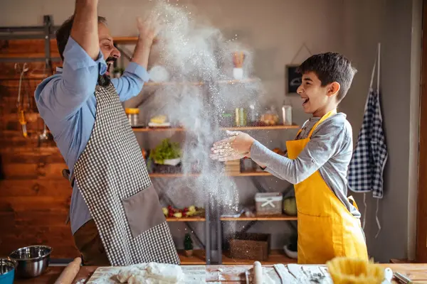 Mann og gutt kaster mel i lufta mens de baker og ler. Foto til artikkel om opplevelsesgaver til farsdag