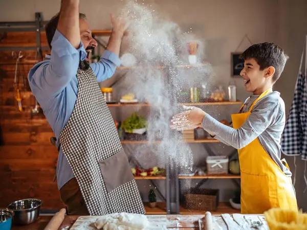 Mann og gutt kaster mel i lufta mens de baker og ler. Foto til artikkel om opplevelsesgaver til farsdag