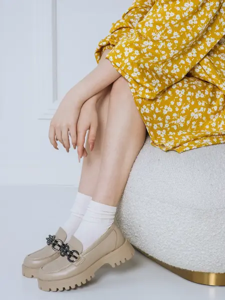 Nærbilde av kvinneben med beige loafers og hvite sokker sittende på en hvit puff med gul og hvitblomstrete kjole