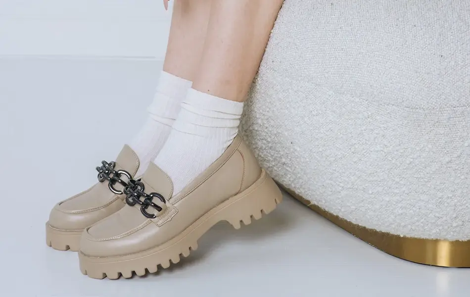 Nærbilde av kvinneben med beige loafers og hvite sokker sittende på en hvit puff med gul og hvitblomstrete kjole