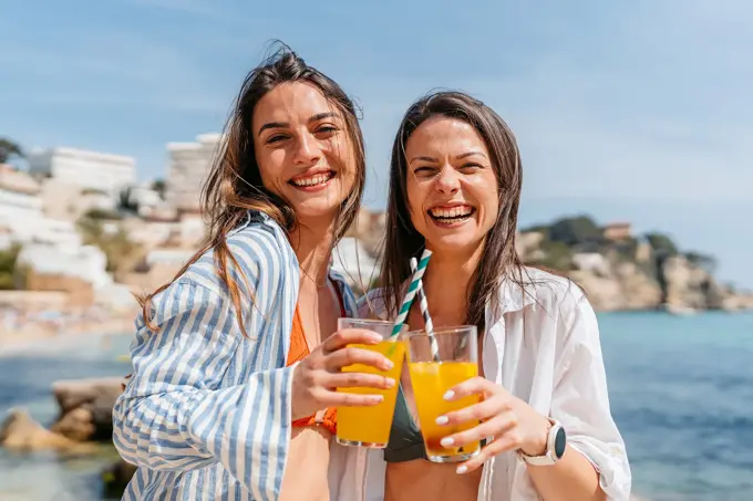 To damer med linskjorte står på strand med lemonade i hånden og smiler til kamera