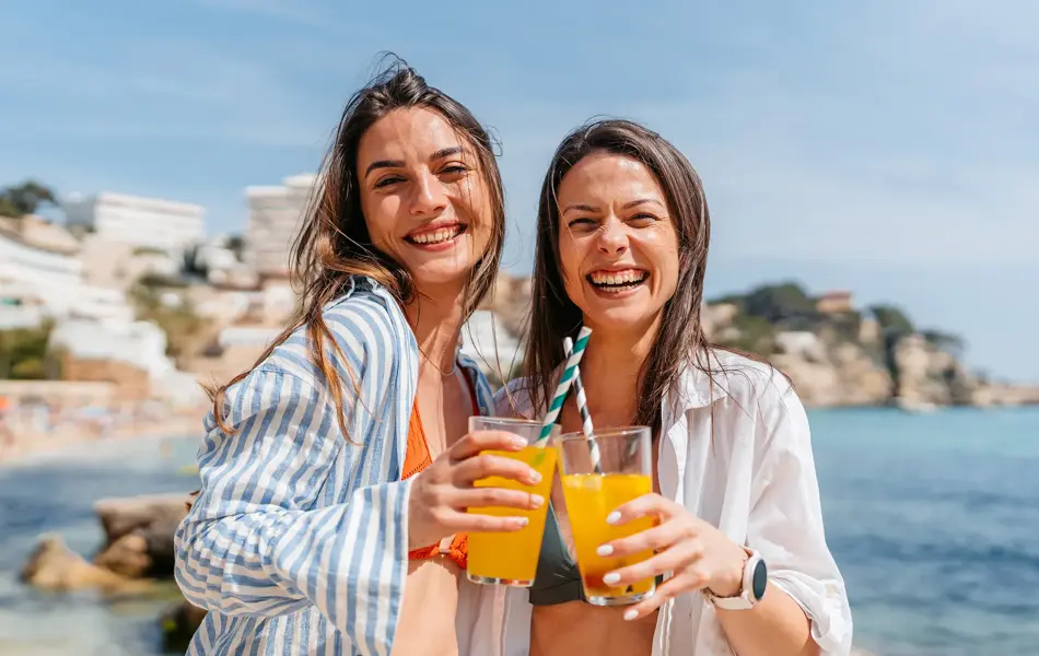 To damer med linskjorte står på strand med lemonade i hånden og smiler til kamera