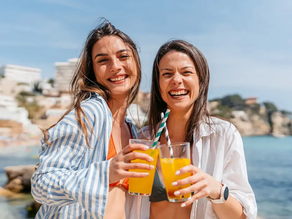 To damer med linskjorte står på strand med lemonade i hånden og smiler til kamera