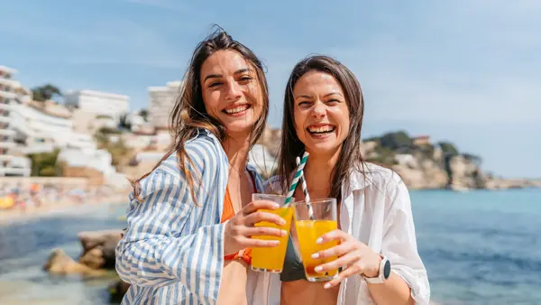 To damer med linskjorte står på strand med lemonade i hånden og smiler til kamera