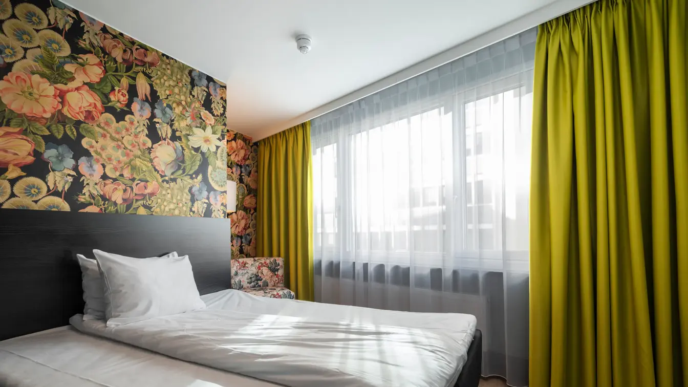 Enkeltrom med enkeltseng, stort vindu, gule gardiner og fargerik tapet. 