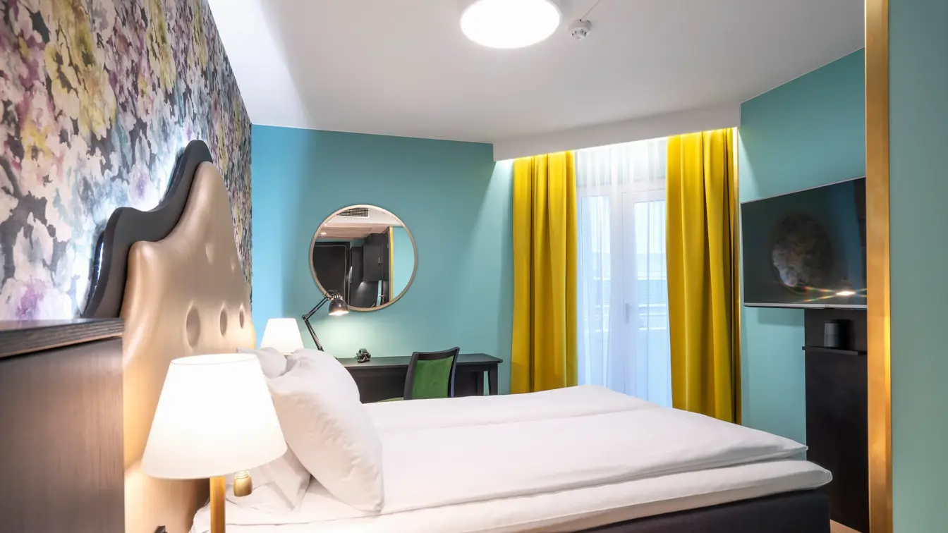 Blomstret tapet bak queen-seng i queen room med turkise farger og gule gardiner, tv og arbeidsplass på Thon Hotel Cecil i Oslo sentrum