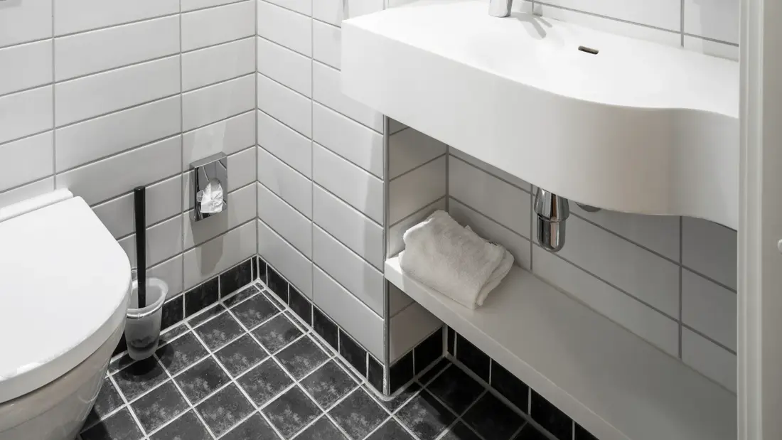 Svarte gulvfliser, hvite veggfliser, toalett, vask, rundt speil.