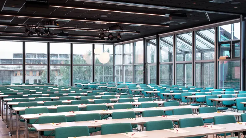 Konferansehall med skrivebord og grønne stoler. Vinduer fra gulv til tak dekker hele rommet.