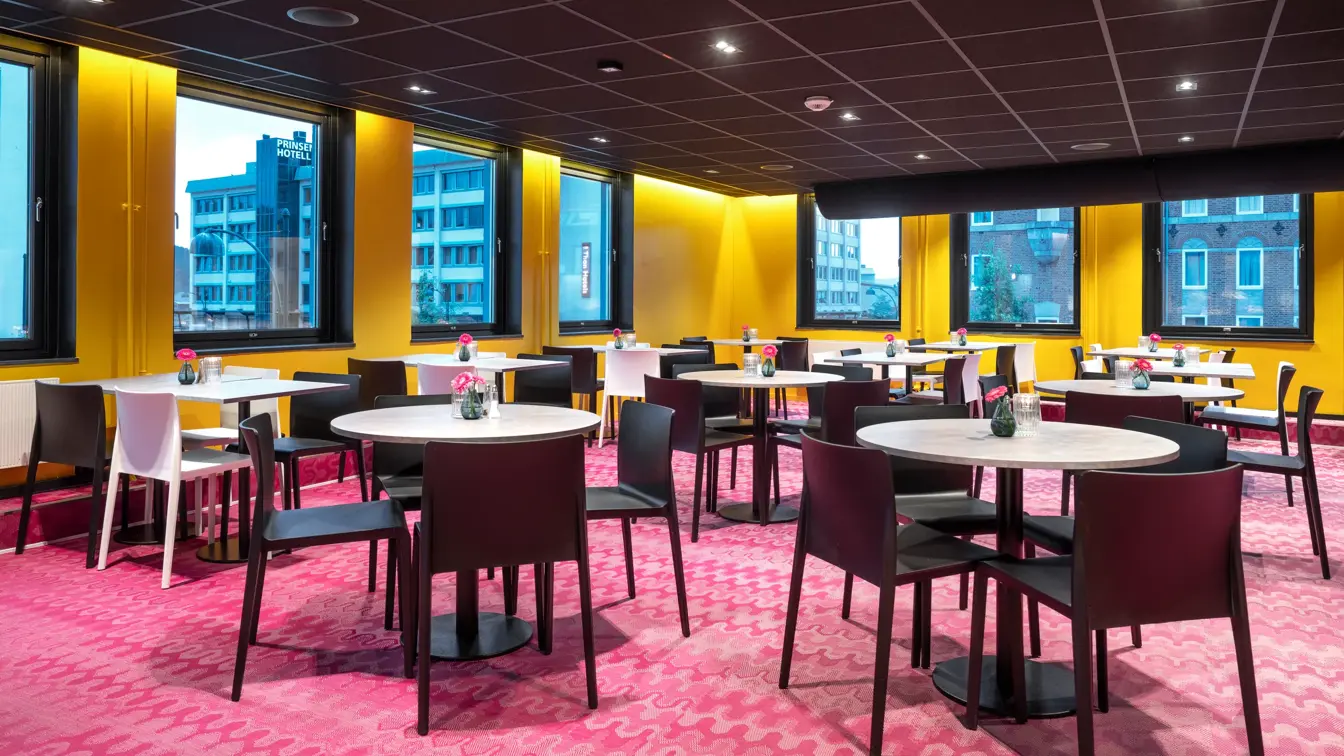 Frokostsal, diverse runde spisebord med svarte stoler, rosa gulvteppe, vinduer, gule vegger. 