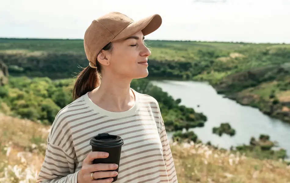 Dame med hvit- og blåstripete genser, mørk beige caps og kaffekopp i hånden nyter solen foran skog- og vannlandskap 
