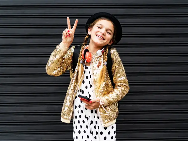 Portrait of happy girl with smartphone wearing hat and golden sequin jacket showing victory sign