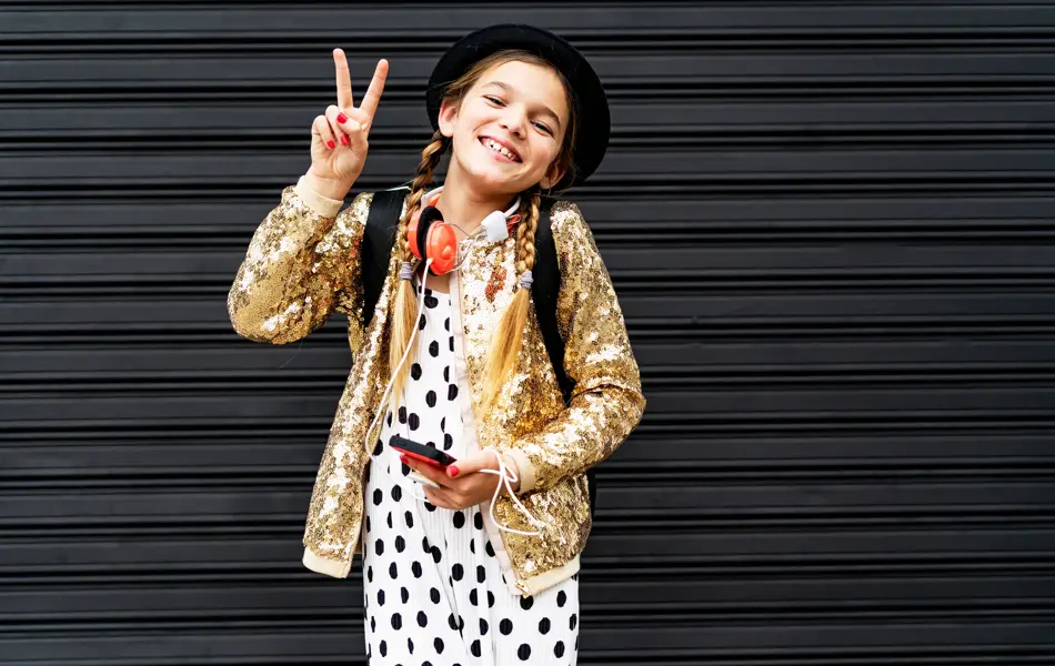 Portrait of happy girl with smartphone wearing hat and golden sequin jacket showing victory sign