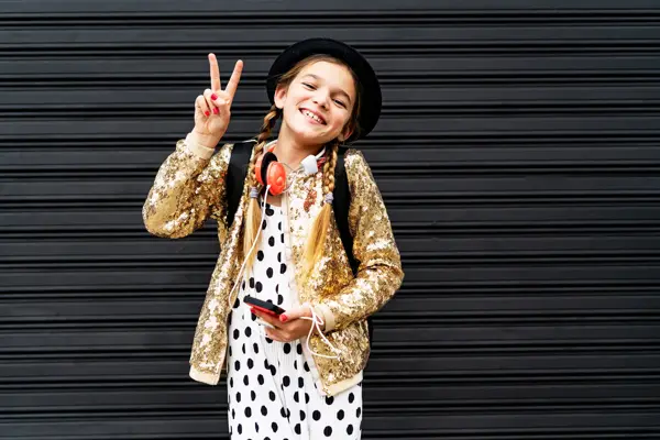 Portrait of happy girl with smartphone wearing hat and golden sequin jacket showing victory sign