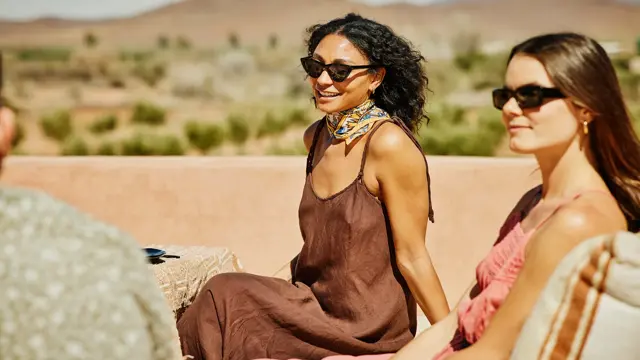 Dame med brun kjole og solbriller sitter på takterrasse og smiler med venner rundt seg 