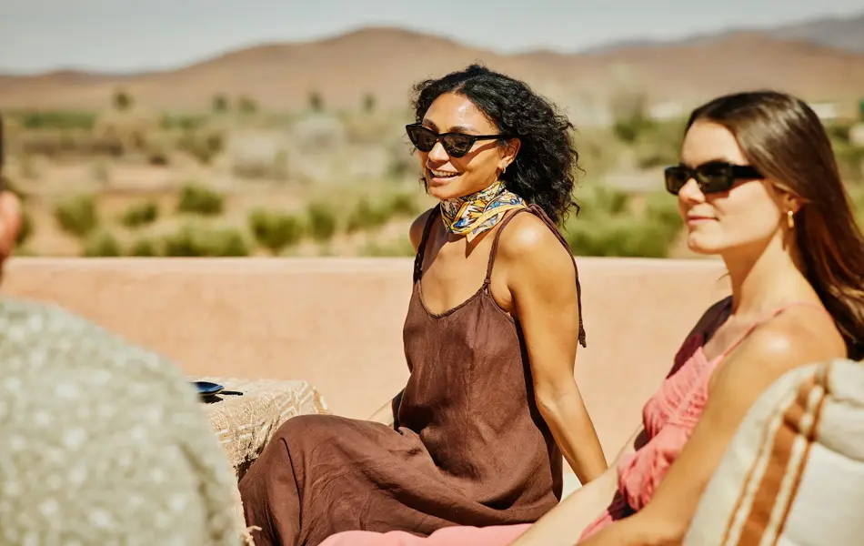 Dame med brun kjole og solbriller sitter på takterrasse og smiler med venner rundt seg 