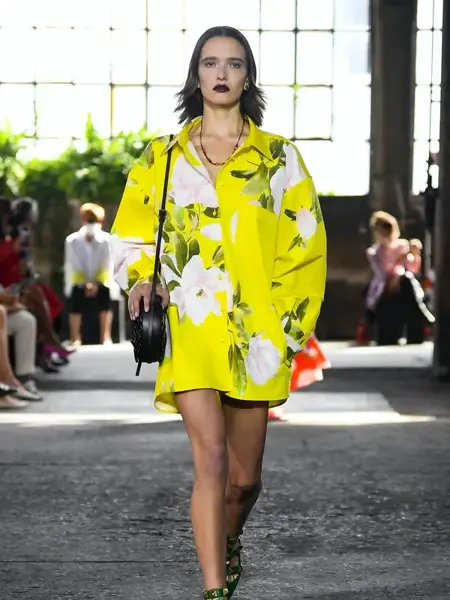 Dame som går på catwalken med en gul kort kjole med store hvite og grønne blomster ppå