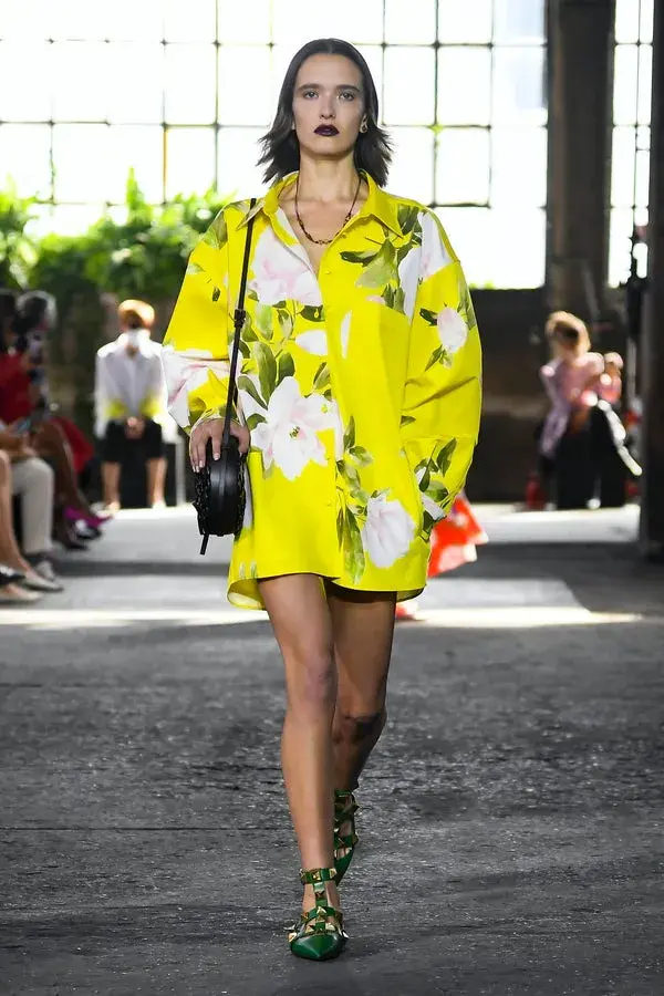 Dame som går på catwalken med en gul kort kjole med store hvite og grønne blomster ppå