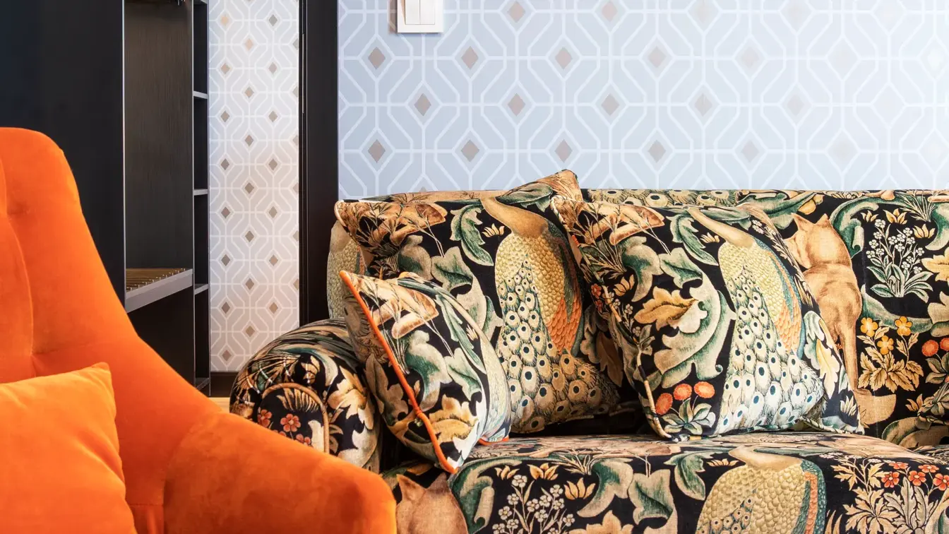 Sittegruppe med oransje stol, mønstret fargerik sofa, mønstret sølvtapet og stort speil i stue i junior suite på Thon Hotel Cecil