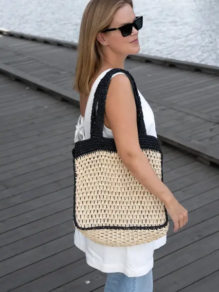 Dame står på brygge med hvit kjole, sorte solbriller og en sort og naturfarget heklet veske i strå.