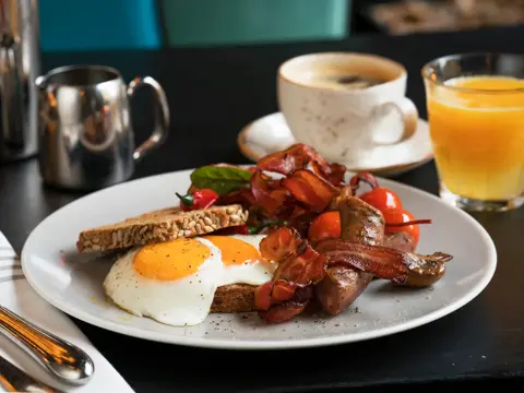 Frokosttallerken med bacon, speilegg, tomater og brødskive. Sammen med en koppkaffe og et glas ferskpresset apelsinjuice