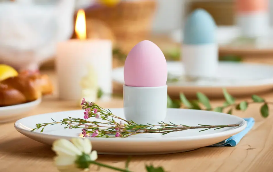 Close-up shot of plates beautifully decorated with colorful Easter eggs in cups and field flowers on blurred background of picturesque decor attributes
