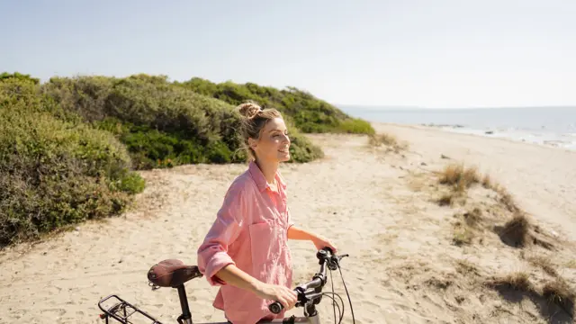 Dame med rosa skjorte holder sykkel på stranden 