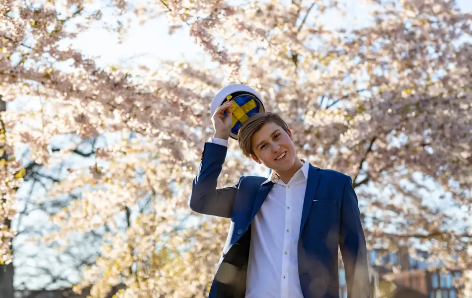 En tenåringsgutt i dress og en svensk studentlue. Han står under kirsebærtrær.