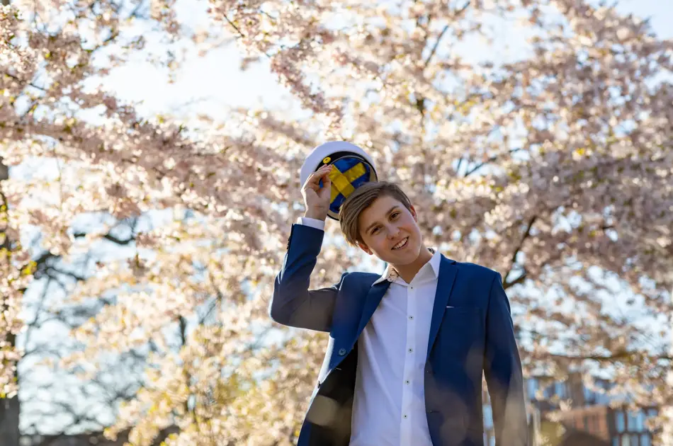 En tenåringsgutt i dress og en svensk studentlue. Han står under kirsebærtrær.