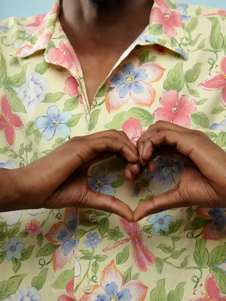 Nærbilde av en mannlig overkropp iført en blomstrete skjorte. Mannen former hendene til et hjerte foran brystet.