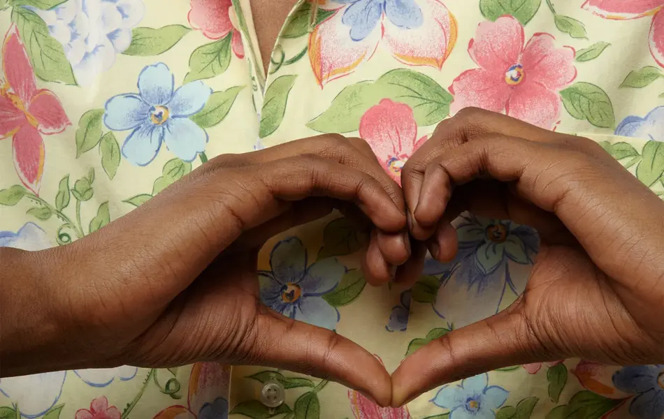 Nærbilde av en mannlig overkropp iført en blomstrete skjorte. Mannen former hendene til et hjerte foran brystet.