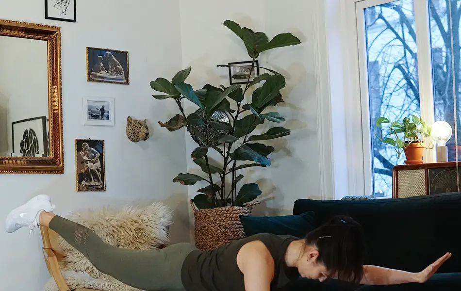 treningsekspert Anna Johansen trener på matte på gulvet, mørkegrønn sofa i bakgrunnen, lys stol med pels oppi, str grønn plante, bilder på veggen