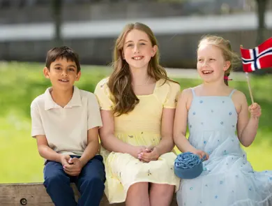 Gutt i hvit piquetskjorte, blå bukse, jente i gul kjole, jente i bå kjole sitter ved siden av hverandre ute.